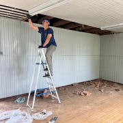 Em uma sala de madeira com parte do forro retirado, um homem de capacete de obra sobe em uma escada.