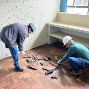 Dentro de uma sala, dois homens de capacete de obra arrancam parquês do chão.