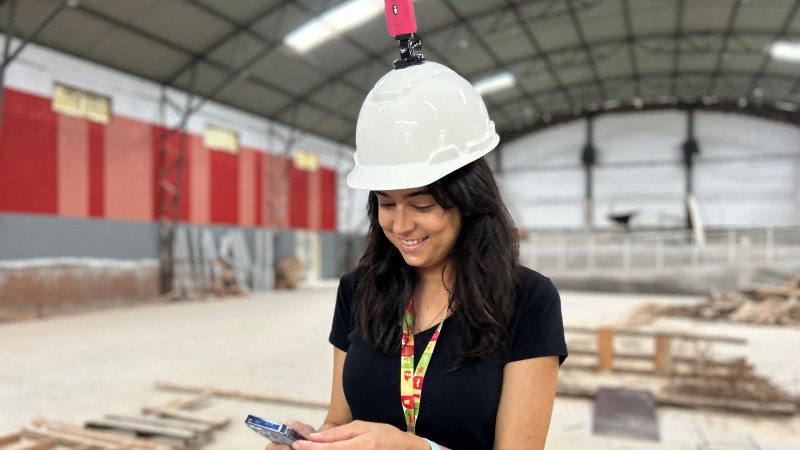 Foto de uma mulher com capacete de obra mexendo no celular em uma obra em um gin&aacute;sio. H&aacute; uma c&acirc;mera rosa sobre o capacete. Ela veste preto, usa um tirante colorido no pesco&ccedil;o e um smartwatch no bra&ccedil;o. Na obra, atr&aacute;s dela, pode-se ver madeiras no ch&atilde;o.