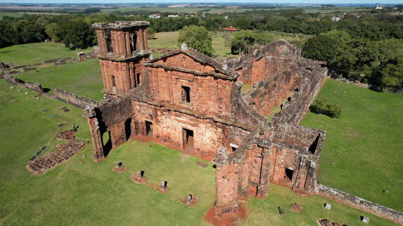 Fotografia a&eacute;rea da Igreja de S&atilde;o Miguel Arcanjo, localizada em S&atilde;o Miguel das Miss&otilde;es, no Rio Grande do Sul. &Eacute; uma constru&ccedil;&atilde;o hist&oacute;rica em ru&iacute;nas feita de pedras avermelhadas, localizada em uma &aacute;rea ampla e aberta coberta por grama verde. A estrutura principal apresenta uma fachada parcialmente preservada, com paredes altas, janelas sem vidro e uma torre retangular &agrave; esquerda, com aberturas em arco. Parte do edif&iacute;cio n&atilde;o possui telhado, permitindo ver o interior vazio e as paredes internas desgastadas pelo tempo.
Ao redor da constru&ccedil;&atilde;o, h&aacute; vest&iacute;gios de outras estruturas menores tamb&eacute;m em ru&iacute;nas, espalhadas pelo terreno. O espa&ccedil;o &eacute; cercado por &aacute;reas verdes e &aacute;rvores, formando um contraste entre o vermelho das pedras e o verde da vegeta&ccedil;&atilde;o. Ao fundo, observa-se uma paisagem rural com campos agr&iacute;colas e algumas edifica&ccedil;&otilde;es distantes, sob um c&eacute;u claro e azul.