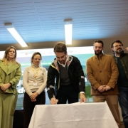 Em um ambiente interno de eventos, seis pessoas est&atilde;o posicionadas diante de um painel de fundo. No centro da cena, uma pessoa inclinada sobre uma mesa coberta por uma toalha branca assina um documento com uma caneta. As demais permanecem em p&eacute; ao redor, acompanhando o ato.
&Agrave; esquerda da imagem, duas pessoas usam roupas escuras e uma terceira veste um conjunto verde-claro. Pr&oacute;xima ao centro, outra pessoa usa blusa clara e crach&aacute; pendurado no pesco&ccedil;o. &Agrave; direita, duas pessoas vestem casacos em tons escuros e terrosos.
Ao fundo, h&aacute; um grande painel de divulga&ccedil;&atilde;o nas cores verde e branco, com o texto &ldquo;RS Seguro&rdquo; vis&iacute;vel na parte superior. No lado direito do painel aparece a fotografia de uma pessoa sorridente. O teto possui lumin&aacute;rias retangulares compridas, que iluminam uniformemente o espa&ccedil;o.
A imagem registra um momento formal de assinatura de documento durante um evento institucional, com os participantes reunidos em volta da mesa acompanhando a cerim&ocirc;nia.