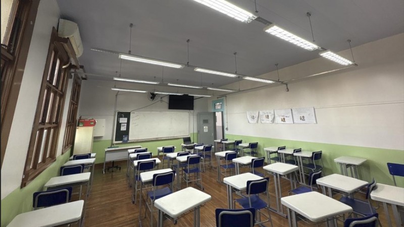 Fotografia de uma sala de aula vazia. O ambiente &eacute; amplo e bem iluminado, com piso de madeira e paredes pintadas em dois tons: verde-claro na parte inferior e branco na superior. No lado esquerdo, h&aacute; uma sequ&ecirc;ncia de janelas de madeira com vidros, permitindo a entrada de luz natural. Acima das janelas, v&ecirc;-se um aparelho de ar-condicionado.
No centro e &agrave; direita da sala, est&atilde;o distribu&iacute;das diversas carteiras escolares individuais, com estrutura met&aacute;lica cinza, tampo branco e cadeiras azuis, organizadas em fileiras voltadas para a frente da sala. Ao fundo, h&aacute; um quadro branco fixado na parede, uma mesa do professor com computador port&aacute;til e uma televis&atilde;o de tela plana instalada acima do quadro. Pr&oacute;ximo ao quadro, h&aacute; uma porta cinza.
O teto &eacute; branco e possui v&aacute;rias lumin&aacute;rias fluorescentes suspensas, alinhadas ao longo da sala. Na parede direita, est&atilde;o afixados trabalhos ou cartazes produzidos por estudantes.