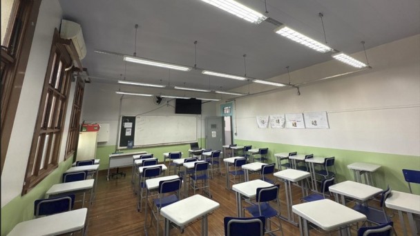 Fotografia de uma sala de aula vazia. O ambiente &eacute; amplo e bem iluminado, com piso de madeira e paredes pintadas em dois tons: verde-claro na parte inferior e branco na superior. No lado esquerdo, h&aacute; uma sequ&ecirc;ncia de janelas de madeira com vidros, permitindo a entrada de luz natural. Acima das janelas, v&ecirc;-se um aparelho de ar-condicionado.
No centro e &agrave; direita da sala, est&atilde;o distribu&iacute;das diversas carteiras escolares individuais, com estrutura met&aacute;lica cinza, tampo branco e cadeiras azuis, organizadas em fileiras voltadas para a frente da sala. Ao fundo, h&aacute; um quadro branco fixado na parede, uma mesa do professor com computador port&aacute;til e uma televis&atilde;o de tela plana instalada acima do quadro. Pr&oacute;ximo ao quadro, h&aacute; uma porta cinza.
O teto &eacute; branco e possui v&aacute;rias lumin&aacute;rias fluorescentes suspensas, alinhadas ao longo da sala. Na parede direita, est&atilde;o afixados trabalhos ou cartazes produzidos por estudantes.
