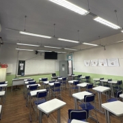 Fotografia de uma sala de aula vazia. O ambiente &eacute; amplo e bem iluminado, com piso de madeira e paredes pintadas em dois tons: verde-claro na parte inferior e branco na superior. No lado esquerdo, h&aacute; uma sequ&ecirc;ncia de janelas de madeira com vidros, permitindo a entrada de luz natural. Acima das janelas, v&ecirc;-se um aparelho de ar-condicionado.
No centro e &agrave; direita da sala, est&atilde;o distribu&iacute;das diversas carteiras escolares individuais, com estrutura met&aacute;lica cinza, tampo branco e cadeiras azuis, organizadas em fileiras voltadas para a frente da sala. Ao fundo, h&aacute; um quadro branco fixado na parede, uma mesa do professor com computador port&aacute;til e uma televis&atilde;o de tela plana instalada acima do quadro. Pr&oacute;ximo ao quadro, h&aacute; uma porta cinza.
O teto &eacute; branco e possui v&aacute;rias lumin&aacute;rias fluorescentes suspensas, alinhadas ao longo da sala. Na parede direita, est&atilde;o afixados trabalhos ou cartazes produzidos por estudantes.