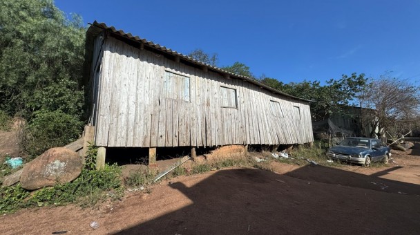 Construção de madeira elevada do chão, com telhado de fibrocimento e paredes desgastadas. Ao redor, há vegetação, terreno de terra e pedras e, à direita, um carro estacionado.