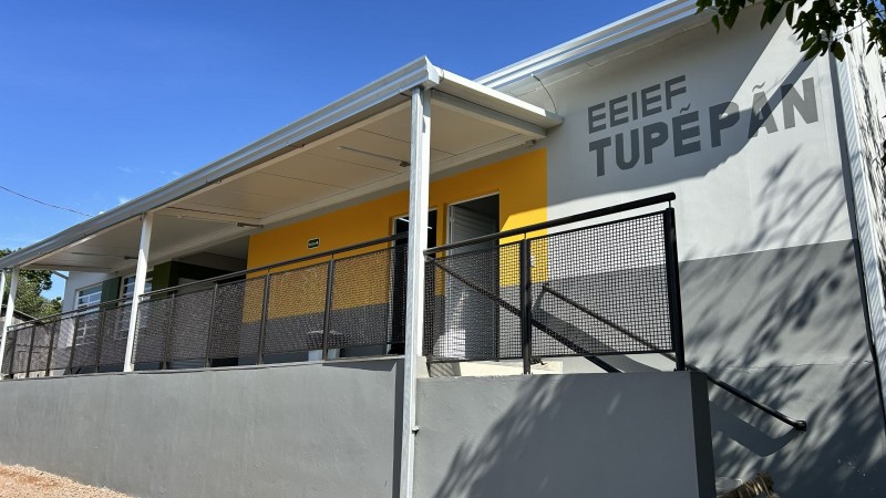 Pr&eacute;dio da EEIEF Tupe Pan em tons de cinza e branco, com detalhes amarelos e verdes. A constru&ccedil;&atilde;o possui corredor coberto, grade met&aacute;lica, escada lateral e &eacute; cercada por &aacute;rvores