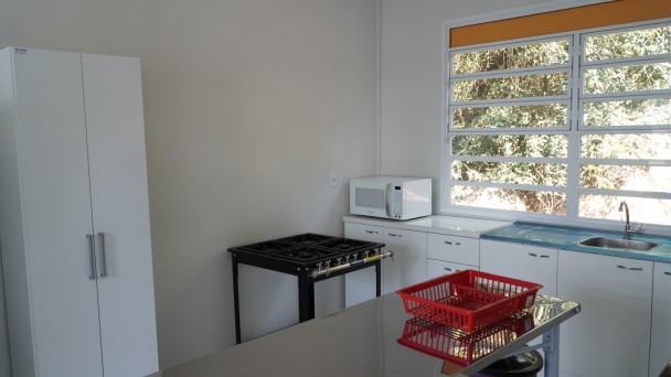 Ambiente de cozinha com paredes claras e mobília predominantemente branca. À esquerda, há um armário alto de duas portas. Ao fundo, uma bancada com armários inferiores abriga um forno micro-ondas branco, uma pia de inox e uma grande janela que permite a entrada de luz natural e vista para a vegetação externa. Próximo à bancada, há um fogão industrial de quatro bocas na cor preta. Em primeiro plano, uma mesa ou bancada com superfície escura possui um cesto plástico vermelho sobre ela. O espaço está organizado, limpo e sem pessoas presentes.