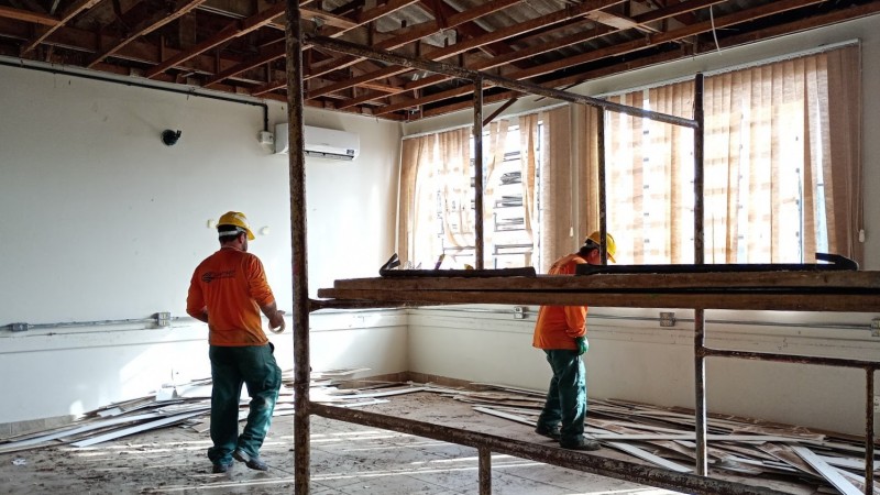 Ambiente interno em reforma, com duas pessoas trabalhadoras usando capacetes amarelos e uniformes nas cores laranja e verde. O teto est&aacute; parcialmente desmontado, expondo vigas e estruturas de madeira. No centro da sala h&aacute; uma estrutura met&aacute;lica de andaime. O piso apresenta sujeira e entulho, com placas e fragmentos de revestimento espalhados pr&oacute;ximo &agrave;s paredes. Grandes janelas na lateral direita permitem a entrada de luz natural, filtrada por persianas verticais claras. Uma unidade de ar-condicionado est&aacute; instalada na parede ao fundo. A imagem registra atividades de obra e adequa&ccedil;&atilde;o em uma sala de escola.