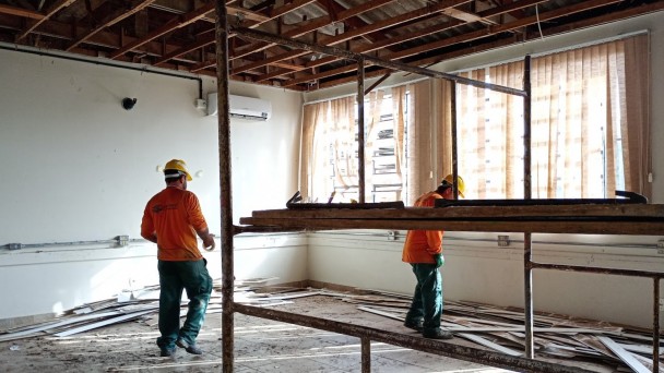 Ambiente interno em reforma, com duas pessoas trabalhadoras usando capacetes amarelos e uniformes nas cores laranja e verde. O teto est&aacute; parcialmente desmontado, expondo vigas e estruturas de madeira. No centro da sala h&aacute; uma estrutura met&aacute;lica de andaime. O piso apresenta sujeira e entulho, com placas e fragmentos de revestimento espalhados pr&oacute;ximo &agrave;s paredes. Grandes janelas na lateral direita permitem a entrada de luz natural, filtrada por persianas verticais claras. Uma unidade de ar-condicionado est&aacute; instalada na parede ao fundo. A imagem registra atividades de obra e adequa&ccedil;&atilde;o em uma sala de escola.