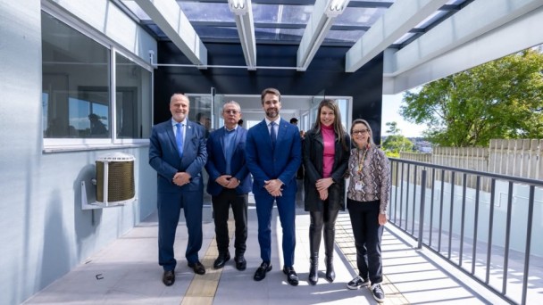 Cinco pessoas posam lado a lado em uma &aacute;rea externa coberta de um pr&eacute;dio. O grupo est&aacute; sobre um piso claro com piso t&aacute;til amarelo ao centro. &Agrave; esquerda, h&aacute; janelas e uma parede cinza; &agrave; direita, um corrim&atilde;o met&aacute;lico e vegeta&ccedil;&atilde;o ao fundo. A cobertura possui estrutura met&aacute;lica e placas transl&uacute;cidas que permitem a entrada de luz natural. As pessoas vestem roupas sociais e est&atilde;o posicionadas de frente para a c&acirc;mera. O ambiente &eacute; bem iluminado e apresenta caracter&iacute;sticas de acessibilidade, como rampa e piso t&aacute;til.