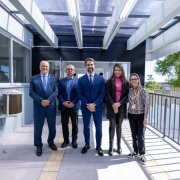 Cinco pessoas posam lado a lado em uma &aacute;rea externa coberta de um pr&eacute;dio. O grupo est&aacute; sobre um piso claro com piso t&aacute;til amarelo ao centro. &Agrave; esquerda, h&aacute; janelas e uma parede cinza; &agrave; direita, um corrim&atilde;o met&aacute;lico e vegeta&ccedil;&atilde;o ao fundo. A cobertura possui estrutura met&aacute;lica e placas transl&uacute;cidas que permitem a entrada de luz natural. As pessoas vestem roupas sociais e est&atilde;o posicionadas de frente para a c&acirc;mera. O ambiente &eacute; bem iluminado e apresenta caracter&iacute;sticas de acessibilidade, como rampa e piso t&aacute;til.