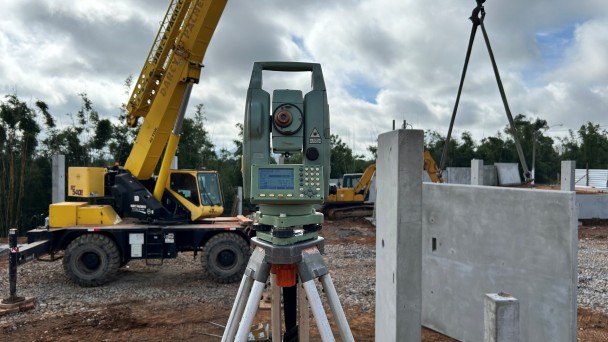 Em um canteiro de obras, um equipamento de topografia montado sobre um trip&eacute; aparece em destaque no centro da imagem. Ao fundo, h&aacute; um guindaste amarelo e uma escavadeira trabalhando na instala&ccedil;&atilde;o de estruturas pr&eacute;-moldadas de concreto. &Agrave; direita, uma grande parede de concreto &eacute; suspensa por cabos para posicionamento. O terreno &eacute; coberto por brita e terra, enquanto &aacute;rvores e vegeta&ccedil;&atilde;o cercam a &aacute;rea ao fundo. O c&eacute;u est&aacute; nublado, com nuvens densas ocupando grande parte da cena.