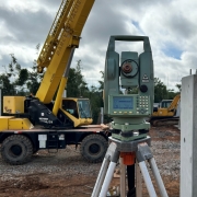 Em um canteiro de obras, um equipamento de topografia montado sobre um trip&eacute; aparece em destaque no centro da imagem. Ao fundo, h&aacute; um guindaste amarelo e uma escavadeira trabalhando na instala&ccedil;&atilde;o de estruturas pr&eacute;-moldadas de concreto. &Agrave; direita, uma grande parede de concreto &eacute; suspensa por cabos para posicionamento. O terreno &eacute; coberto por brita e terra, enquanto &aacute;rvores e vegeta&ccedil;&atilde;o cercam a &aacute;rea ao fundo. O c&eacute;u est&aacute; nublado, com nuvens densas ocupando grande parte da cena.
