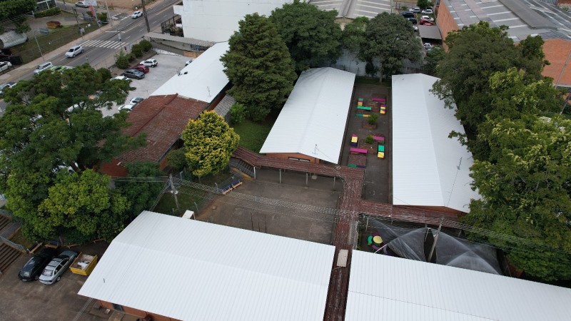 Vista a&eacute;rea de um complexo escolar cercado por &aacute;rvores de grande porte em &aacute;rea urbana. O conjunto &eacute; formado por v&aacute;rios blocos t&eacute;rreos com telhados met&aacute;licos brancos e algumas coberturas de telha avermelhada. No centro, h&aacute; um p&aacute;tio interno pavimentado com bancos e mesas coloridos distribu&iacute;dos ao longo do espa&ccedil;o. Ao redor da escola, aparecem ruas com circula&ccedil;&atilde;o de ve&iacute;culos, estacionamentos, pr&eacute;dios comerciais e residenciais. A vegeta&ccedil;&atilde;o abundante ocupa boa parte do terreno, contrastando com as constru&ccedil;&otilde;es e o entorno urbano. O registro foi feito de cima, oferecendo uma vis&atilde;o ampla da escola e da vizinhan&ccedil;a.