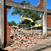Máquina escavadeira amarela realiza a demolição de uma estrutura de alvenaria e concreto. No primeiro plano, há grande quantidade de entulho formado por tijolos e fragmentos de construção espalhados pelo chão. Restam apenas partes das paredes de tijolos vermelhos e pilares de concreto do prédio. Ao fundo, árvores verdes contrastam com o céu azul sem nuvens. A imagem registra uma etapa de demolição de edificação em área escolar.