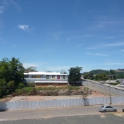 Vista ampla do terreno onde existia um ginásio escolar, após a conclusão da demolição. A área aparece cercada por tapumes metálicos cinza e apresenta solo nivelado, montes de entulho e restos de vegetação removida. Ao fundo, está o prédio da escola, de dois pavimentos, pintado em tons claros, cercado por árvores. Há ruas pavimentadas, postes de energia e alguns veículos ao redor do terreno. O céu azul com poucas nuvens completa a paisagem urbana e rural ao redor da escola.
