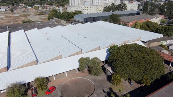 Vista a&eacute;rea de um grande pr&eacute;dio com cobertura met&aacute;lica branca rec&eacute;m-instalada. O telhado ocupa quase toda a imagem e apresenta superf&iacute;cies amplas e uniformes. Na parte inferior, h&aacute; uma &aacute;rea pavimentada com formato circular, &aacute;rvores de grande porte e dois ve&iacute;culos estacionados. Ao redor do edif&iacute;cio, observam-se outras constru&ccedil;&otilde;es, ruas e &aacute;reas urbanizadas. A fotografia destaca a conclus&atilde;o da recupera&ccedil;&atilde;o da cobertura da edifica&ccedil;&atilde;o e sua integra&ccedil;&atilde;o com o entorno.