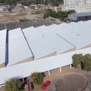 Vista a&eacute;rea de um grande pr&eacute;dio com cobertura met&aacute;lica branca rec&eacute;m-instalada. O telhado ocupa quase toda a imagem e apresenta superf&iacute;cies amplas e uniformes. Na parte inferior, h&aacute; uma &aacute;rea pavimentada com formato circular, &aacute;rvores de grande porte e dois ve&iacute;culos estacionados. Ao redor do edif&iacute;cio, observam-se outras constru&ccedil;&otilde;es, ruas e &aacute;reas urbanizadas. A fotografia destaca a conclus&atilde;o da recupera&ccedil;&atilde;o da cobertura da edifica&ccedil;&atilde;o e sua integra&ccedil;&atilde;o com o entorno.